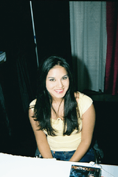 A Black Haired Woman Wearing A Yellow Top And Jeans Sitting Behind A Table Looking 1